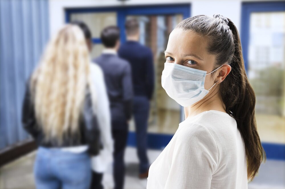 Frau mit Gesichtsmaske wartet in der Schlange vor einer Glastüre